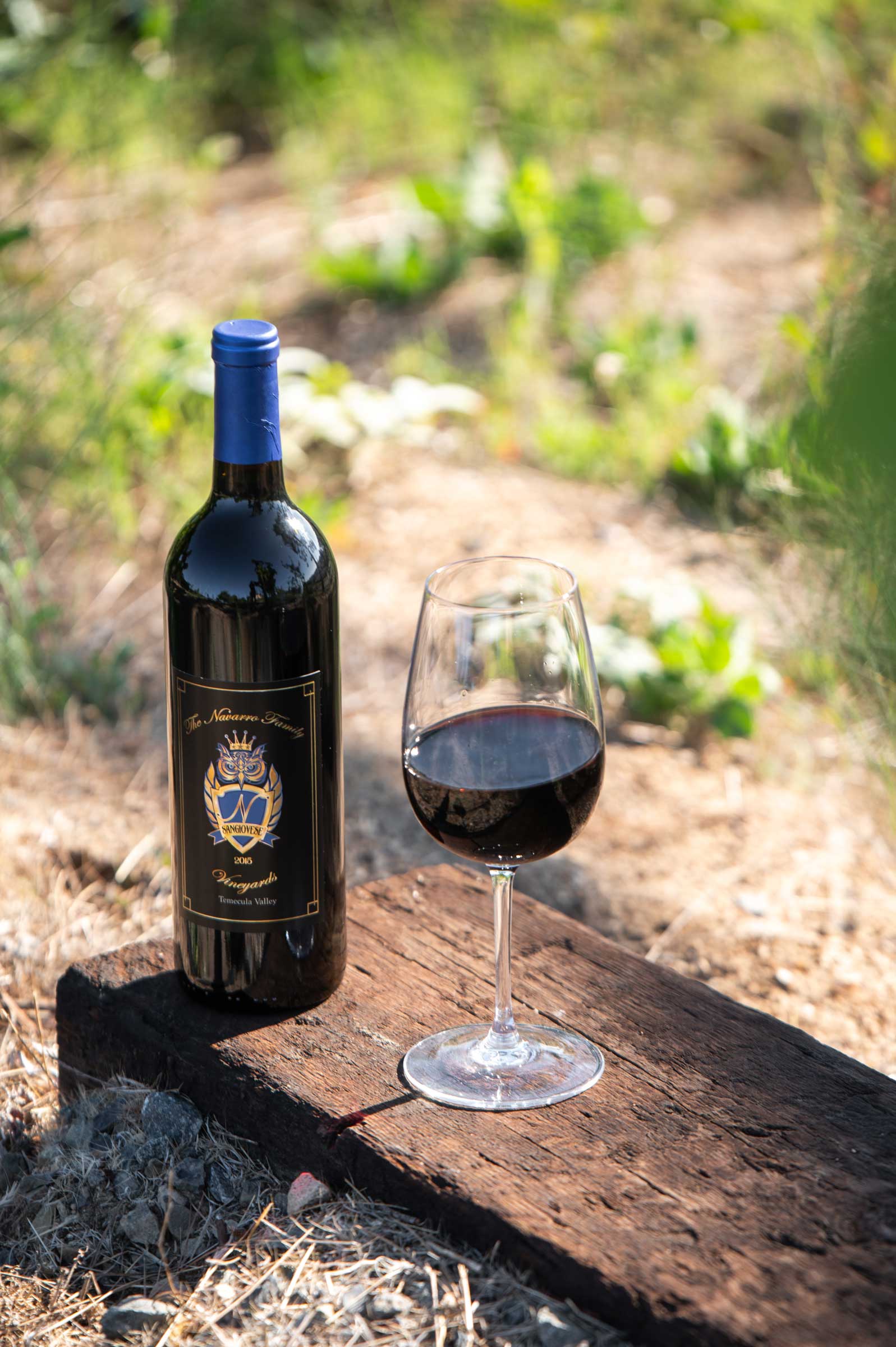 bottle of Navarro Family Vineyards Sangiovese beside a full glass on a wooden bench among the vines